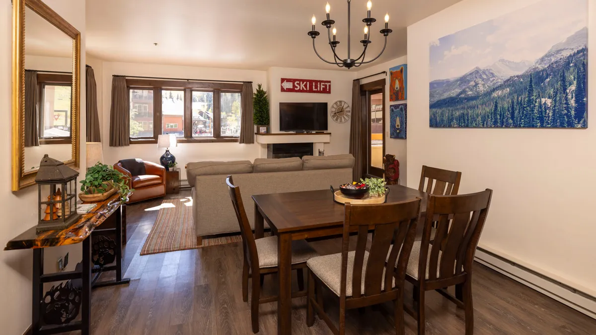 A seamless open-plan space where a dining table with chairs sits adjacent to a comfortable sofa. The area is flooded with natural light from large windows, and the decor includes mountain landscape artwork and a ski lift sign, tying the interior to the mountain setting.
