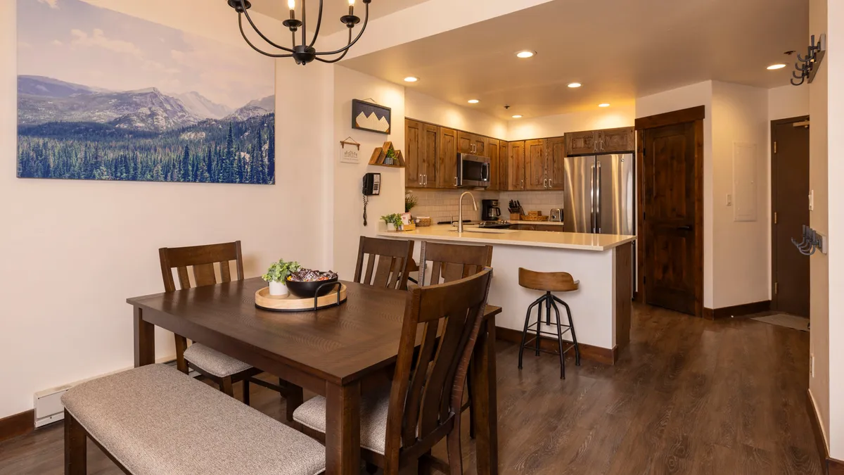An inviting dining area seamlessly connected to a modern kitchen, featuring a large dining table set under a stylish chandelier. The kitchen island and counters are equipped with modern appliances. A mountain landscape painting decorates the wall, and large windows bring in natural light.
