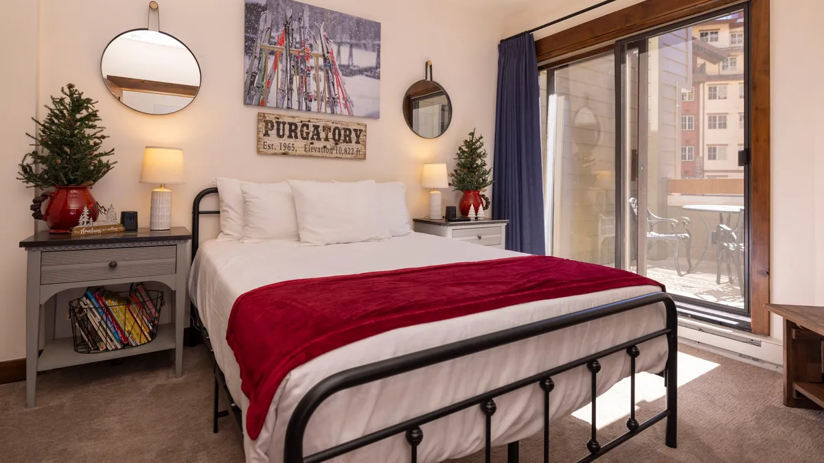 A warm, welcoming bedroom with a bed dressed in a red blanket, plants on the nightstands, and artwork on the wall. A sliding glass door lets in natural light, creating a space perfect for relaxation or enjoying morning views.