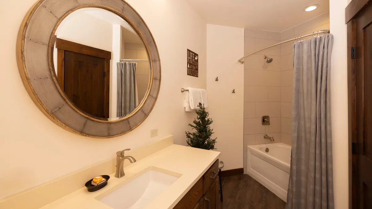 A sparkling clean bathroom featuring a round mirror, vanity with sink, shower curtain, small decorative tree, and wall decor. The mood is tranquil and refreshing, suggesting a space designed for privacy and relaxation.
