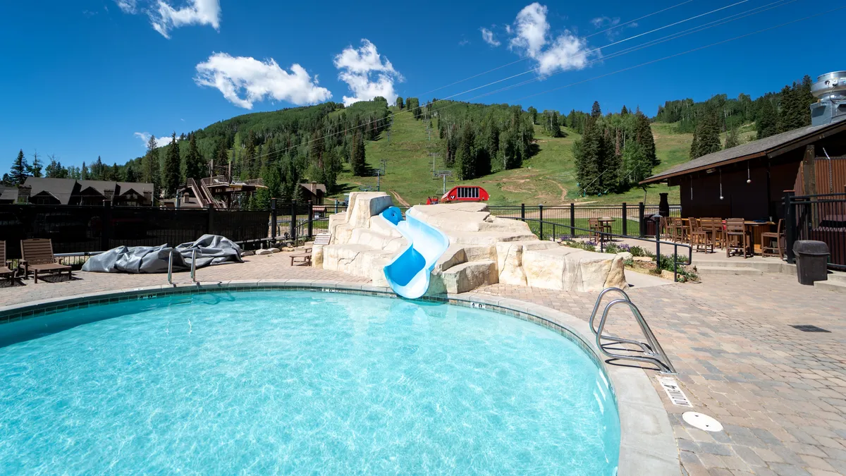 A vibrant outdoor swimming pool area surrounded by a lush green hillside and an inviting outdoor seating section. The pool features a water slide, and there’s ample space for sunbathing and family gatherings, capturing the spirit of summer fun and relaxation at the resort.