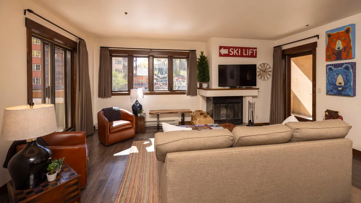 A welcoming living room featuring a large fireplace as the focal point, surrounded by a plush, comfortable sofa, wooden coffee table, and large windows offering scenic mountain views. Art decorates the walls, and the soft lighting enhances the cozy, inviting atmosphere.