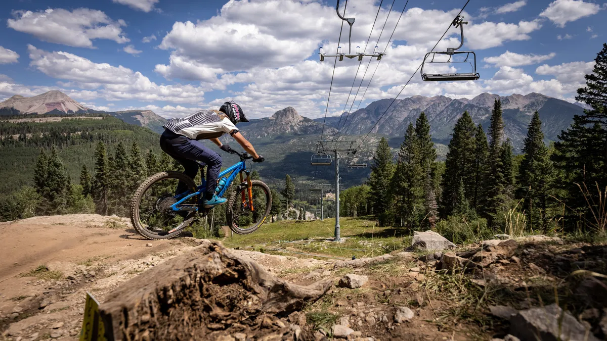 A mountain biker speeds down a lush, green trail, with a dramatic mountain range and ski lift in the background. The environment is vibrant, with summer foliage and the thrill of high-adrenaline recreation.