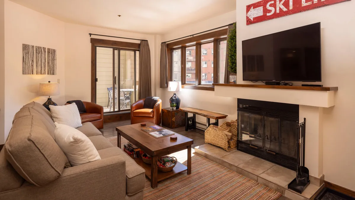 The image showcases a warm, inviting living room featuring a plush sofa set around a glowing fireplace and a mounted TV, with a rustic 'Ski Lift' sign adding mountain charm. The atmosphere is cozy and perfect for relaxing or spending time together after outdoor adventures.
