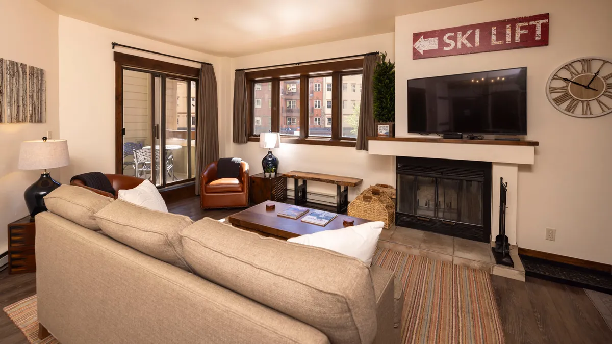 A cozy living room interior with a plush sofa, glowing fireplace, and large windows revealing a hint of the outdoors. A ski lift sign adds a touch of local character, and the space is set for relaxing or gathering after a day out.