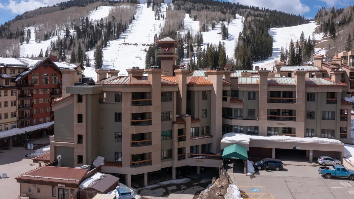 A snow-covered resort building sits at the edge of expansive ski slopes, with dramatic mountain peaks rising in the background. The scene captures people enjoying the outdoors, with hints of après-ski activities and the cozy architecture of the lodging.