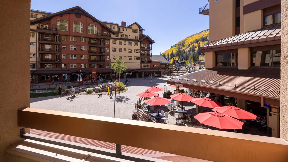 A lively resort plaza with people dining under red umbrellas, surrounded by resort buildings and a stunning mountain backdrop. The vibrant outdoor atmosphere suggests access to local shops, restaurants, and events.