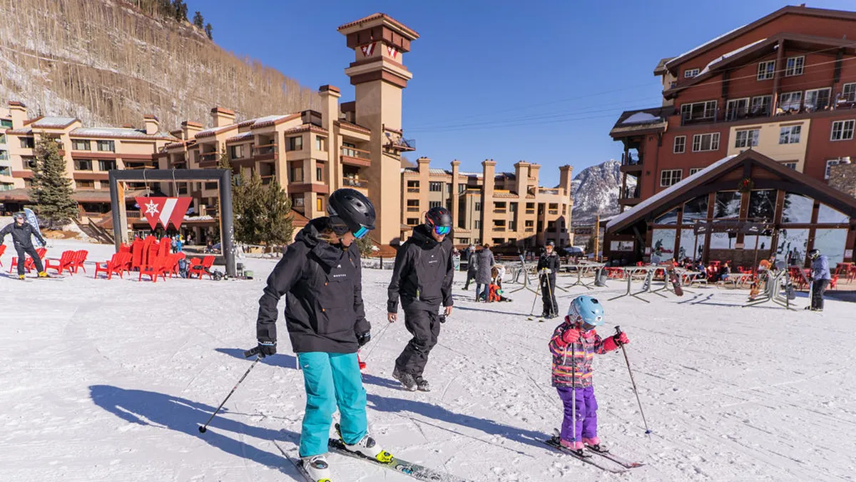 A lively exterior scene featuring families and couples skiing down a snow-covered slope, with charming resort buildings in the background. The image exudes a playful, adventurous mood and showcases the vibrant atmosphere of Purgatory Village.