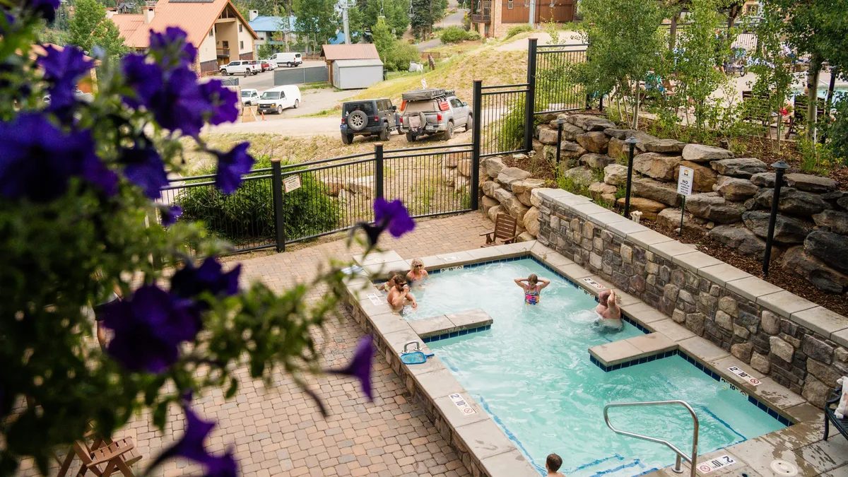 An exterior view of a hot tub surrounded by people enjoying themselves, set amidst lush greenery and colorful flowers. The scene feels casual and social, emphasizing relaxation and opportunities for social gatherings.
