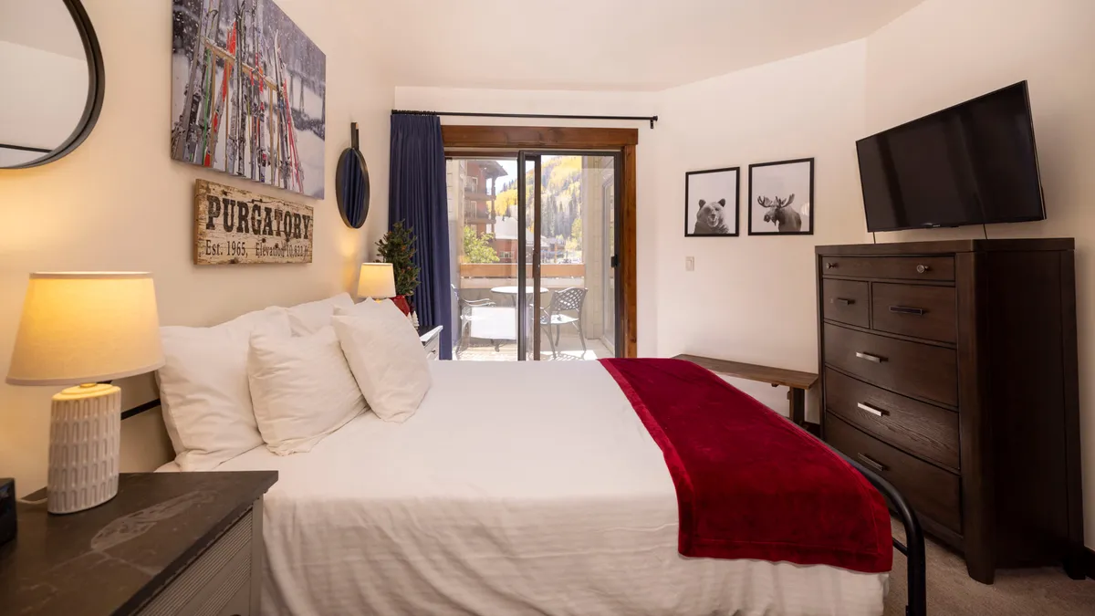 This inviting bedroom features a bed adorned with a red throw, a sliding glass door that opens to the outside, and artful décor. The sliding door hints at access to a private balcony and mountain views, suggesting a seamless indoor-outdoor connection.