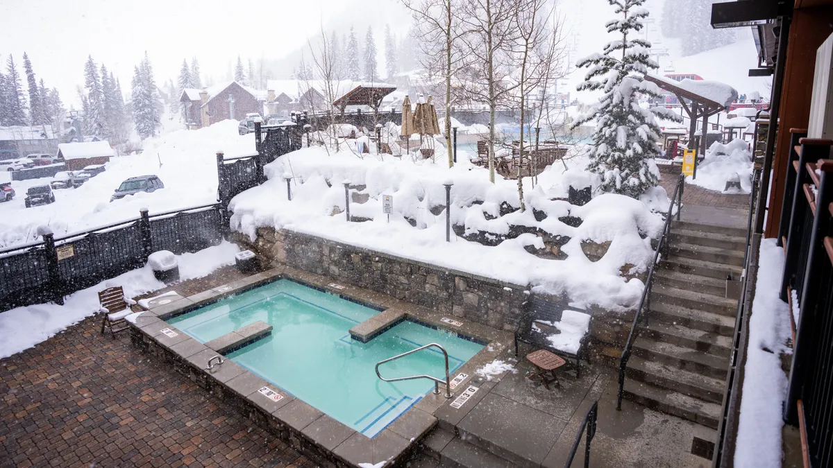 An exterior view of the lodge's bubbling hot tub set in a peaceful, snow-covered landscape surrounded by trees and a stone wall. The scene feels tranquil and welcoming, ideal for families to gather, relax, and soak after exploring the resort or hitting the slopes.