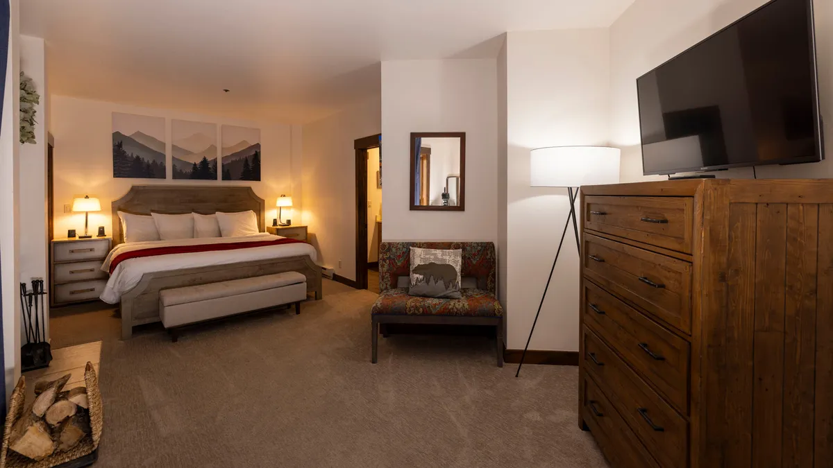 A restful bedroom scene featuring a plush bed, tasteful artwork above the headboard, a dresser, and a glowing lamp. The bedding looks soft and inviting, and the overall decor exudes tranquility and comfort.