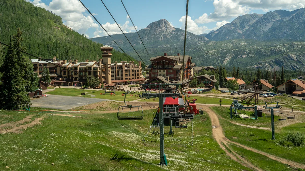 This exterior image captures the essence of resort life, featuring a ski lift, green grass, the lodge, and towering mountains in the background. It suggests year-round activities like skiing, hiking, and mountain biking, with open spaces inviting families to explore and play.