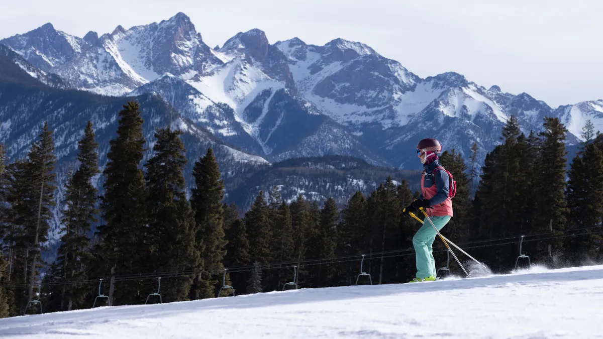 This exterior photograph captures a skier traversing down a slope in front of the majestic, snow-covered San Juan Mountains, fringed by tall pine trees. The crisp mountain air and sweeping views are immediately apparent, immersing viewers in the mountain environment.